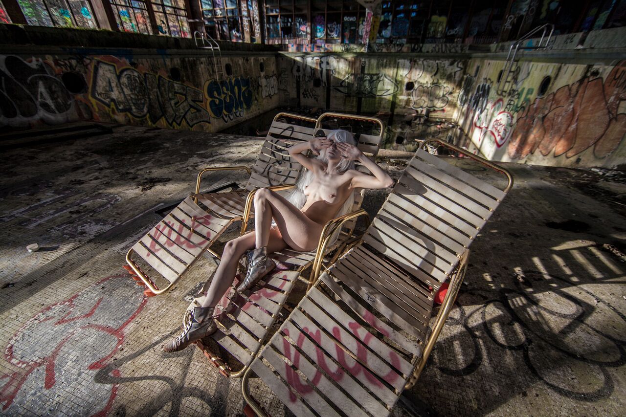 A performer reclines on striped lounge chairs in a drained, graffiti-marked pool.