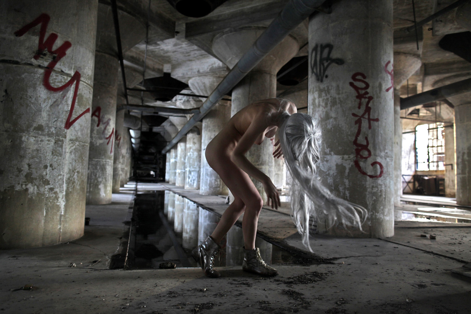 Julia Sinelnikova bends amid broken industrial structures beside water, framed by rusted beams and concrete.