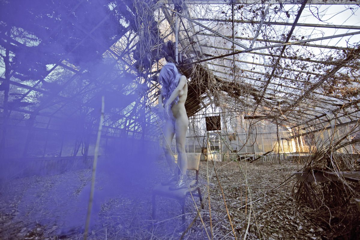Purple smoke moves around a performer in an overgrown, glass-roofed abandoned structure.