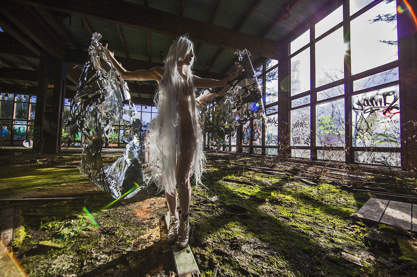 A performer extends reflective, wing-like material inside a sunlit abandoned building.